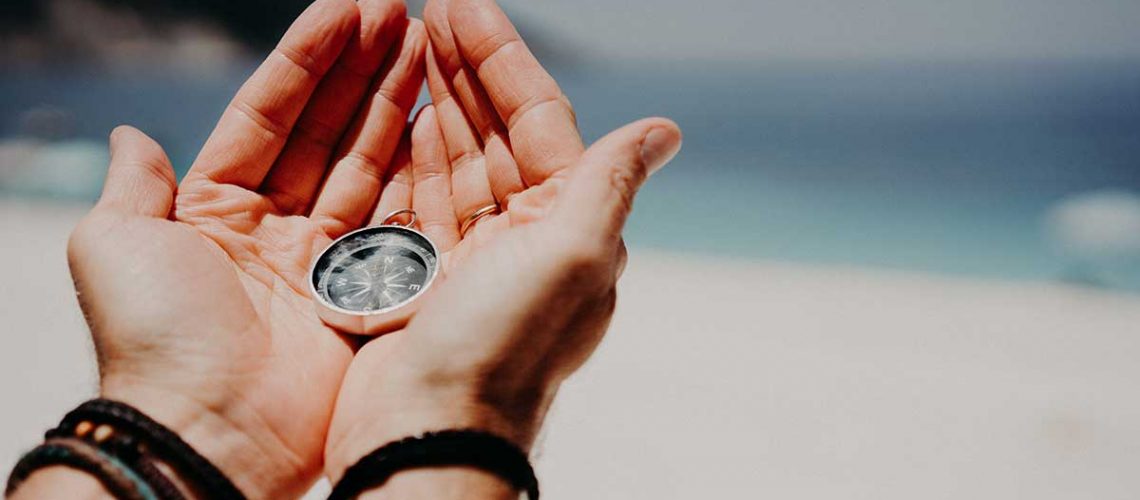 mans-hand-holding-compass-against-the-backdrop-of-beach-and-sea-the-concept-of-travel_1200x628