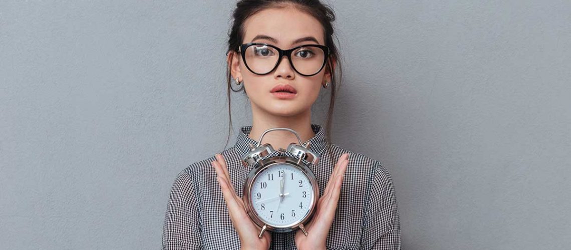 young-surprised-asian-woman-in-glasses-and-shirt-holding-the-clock-in-hands-isolated_1200x628